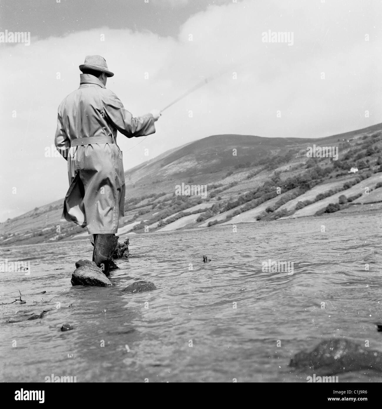 1950, storico, un pescatore che indossa un impermeabile, trampolieri e cappello, in piedi, pesca con la mosca (pesca) per il salmone in un fiume del Galles. Foto Stock