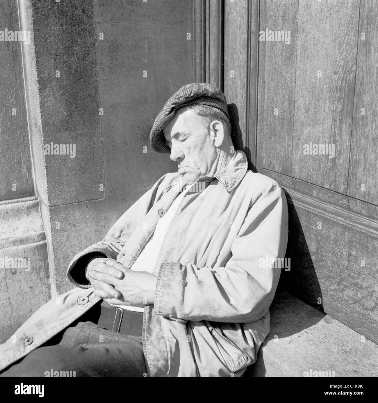 Anni '1950, un vecchio stanco, con un impermeabile e un berretto piatto, forse un vagabondo, fa un pomeriggio a snooze sui gradini di Hungerford House, Embankment, Londra. Foto Stock