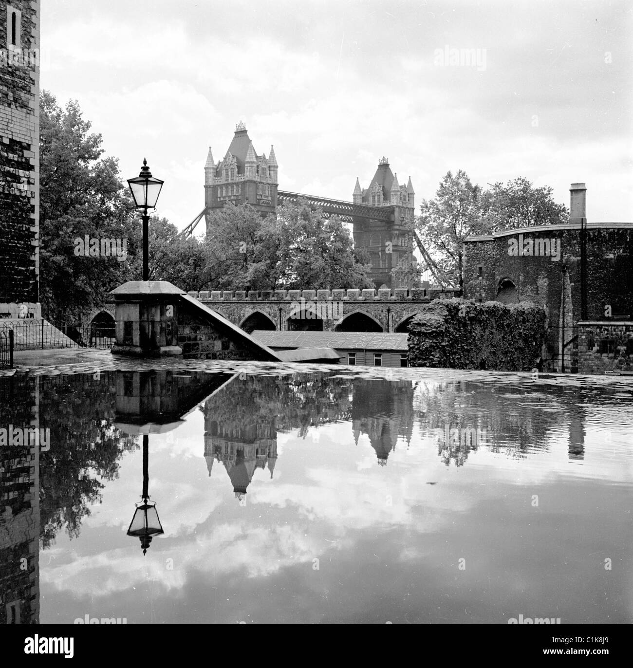 Anni '1950, una piscina d'acqua copre il passaggio acciottolato vicino ai gradini di Tower Green, Londra. Tower Bridge visto in lontananza. Foto Stock