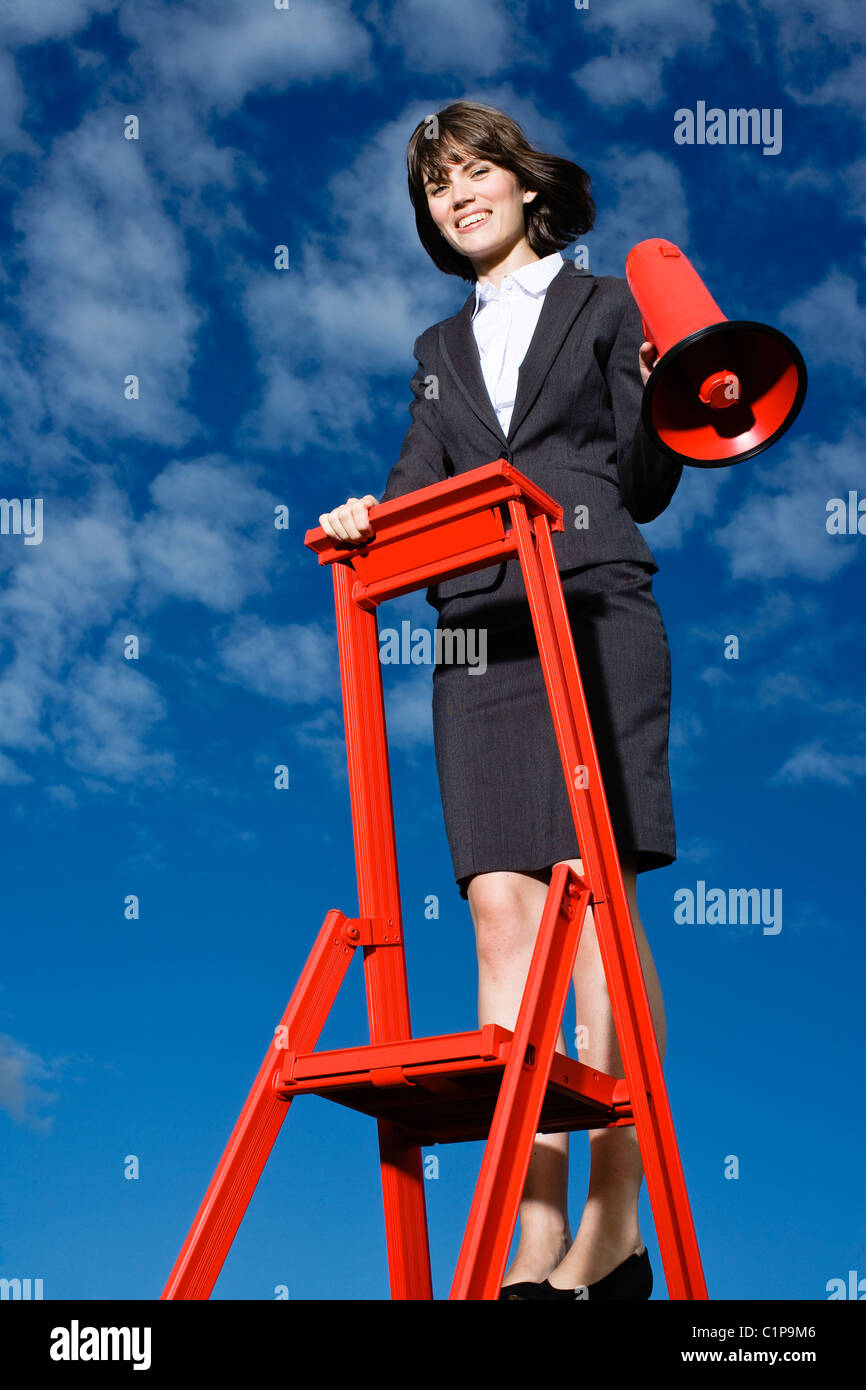 Sorridente imprenditrice permanente sulla parte superiore della scaletta rosso contro il cielo blu e rosso di contenimento megafono Foto Stock