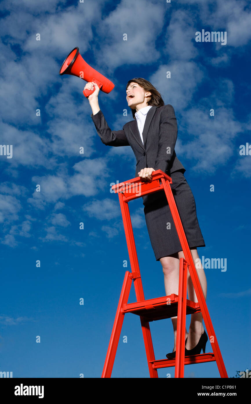 Imprenditrice permanente sulla parte superiore della scaletta rosso contro il cielo blu e urlando attraverso il red megafono Foto Stock
