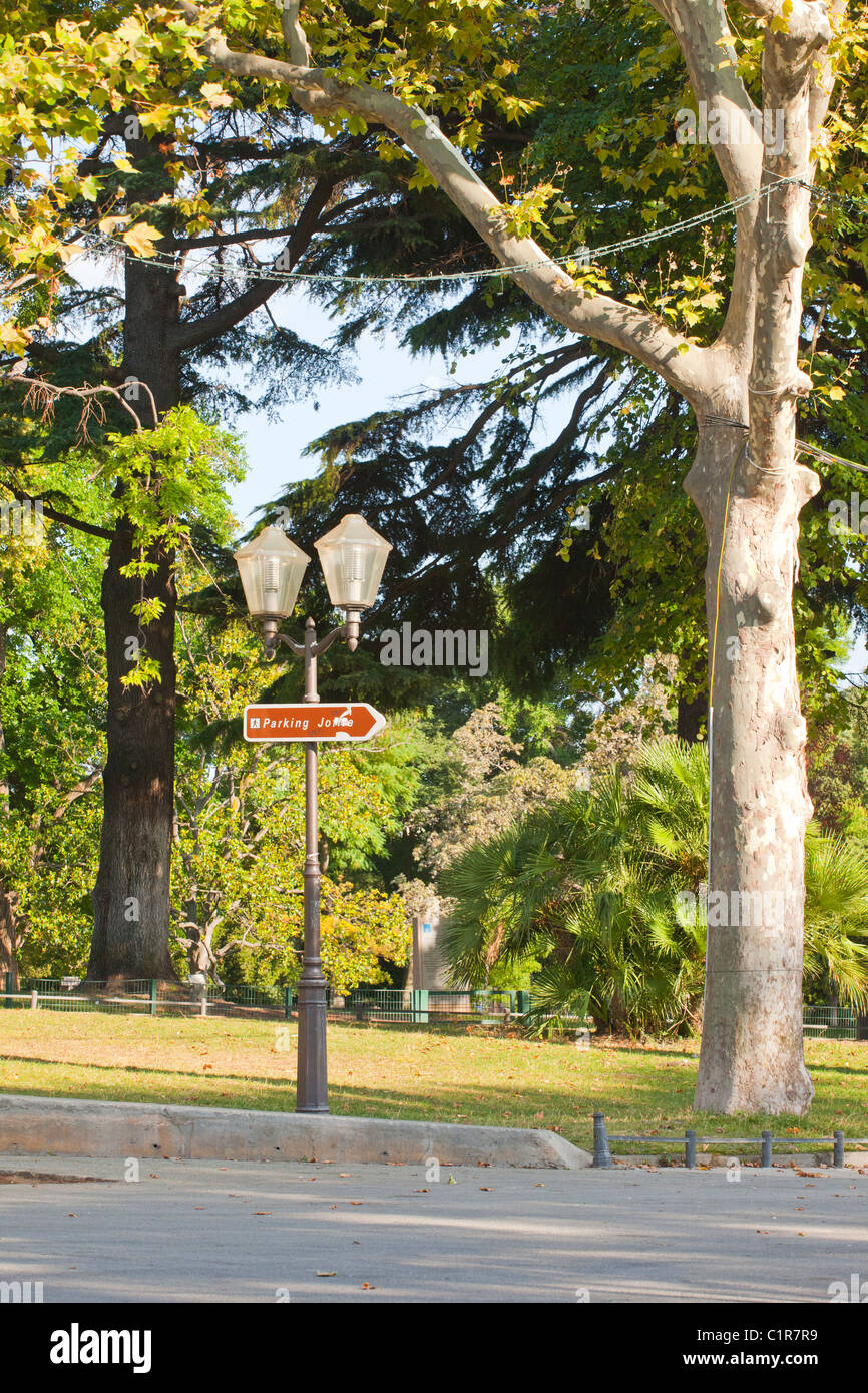 Tranquillo parco di Montpellier con lampione e segnale di parcheggio direzionale tra alberi lussureggianti in una giornata di sole. Foto Stock