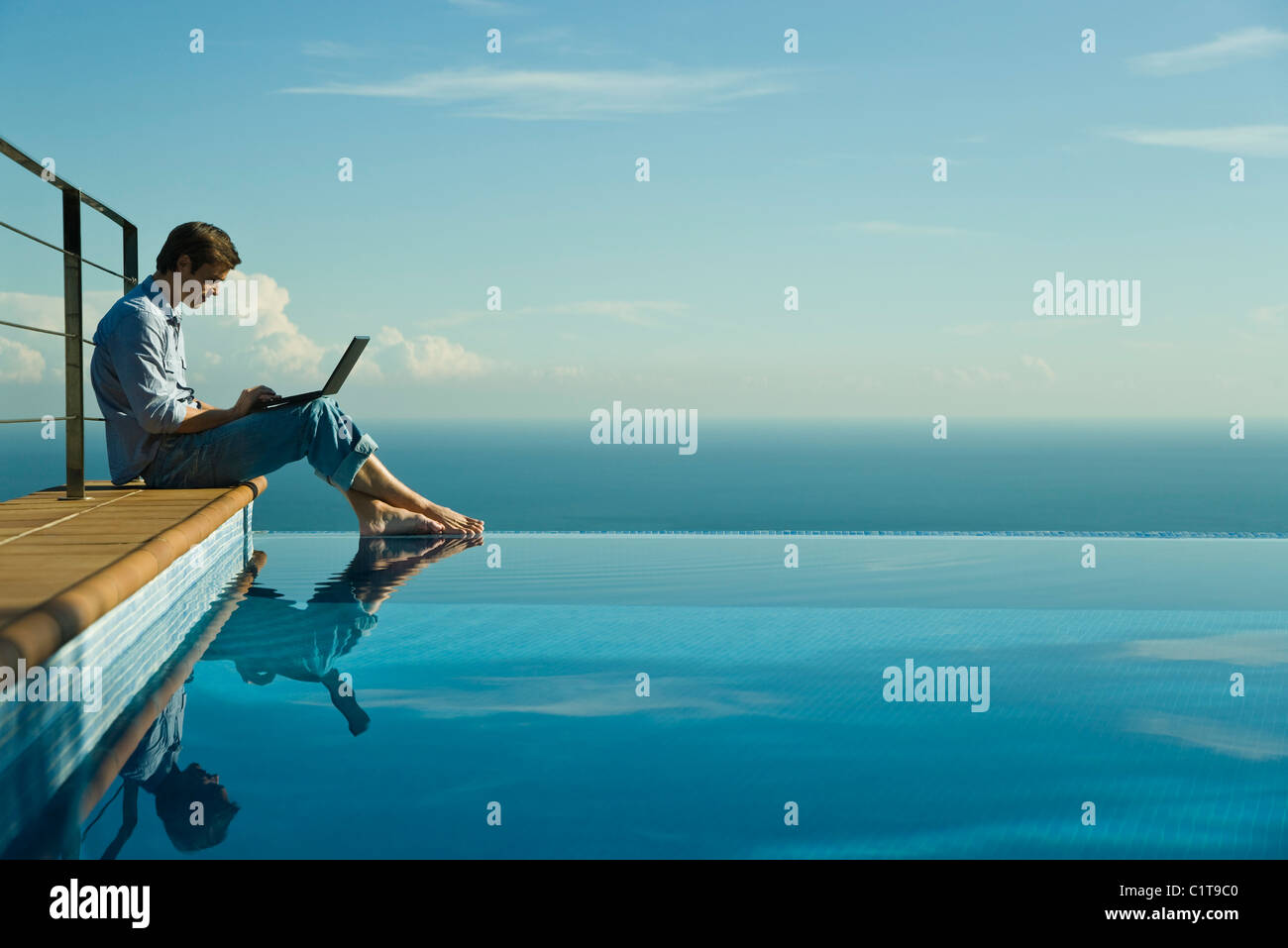 Uomo seduto accanto alla piscina infinity, utilizzando il computer portatile Foto Stock