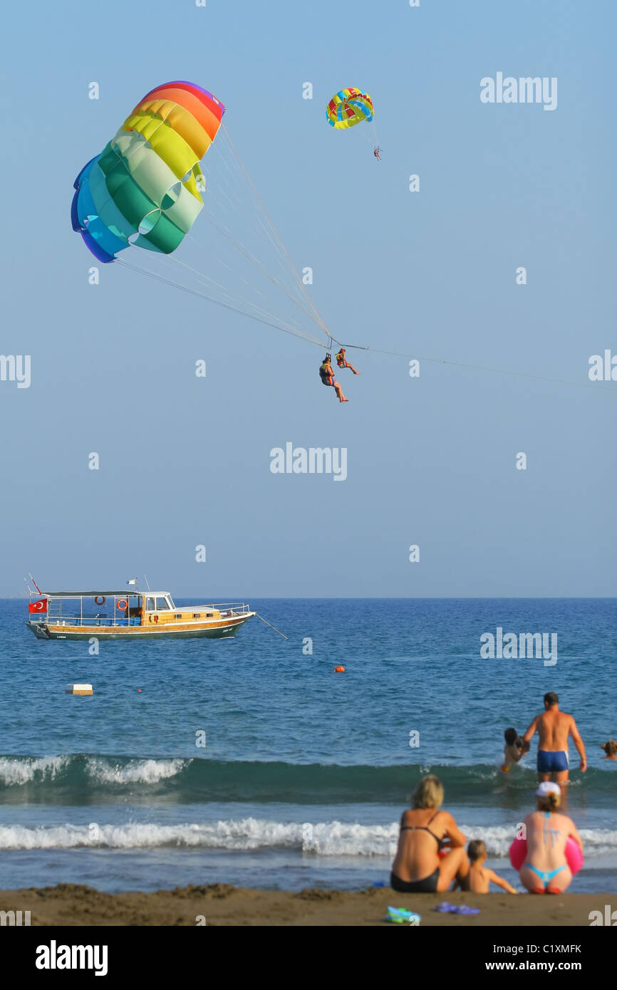 Le persone a rilassarsi sulla spiaggia e trainate dietro una barca sul multi-paracadute colorato. Sarigerme, Mar Egeo, il mare della Turchia Foto Stock