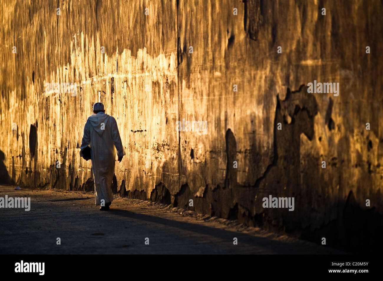 Un uomo arabo si allontana dal visualizzatore giù per una strada di Marrakech al tramonto. La parete dietro l'uomo brilla di un colore oro scuro. Foto Stock