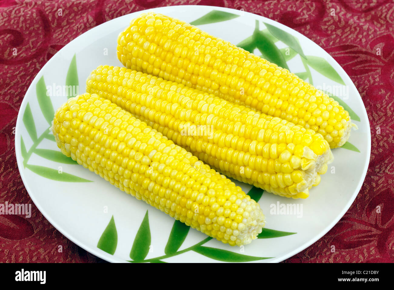 Tre luminoso giallo imburrato orecchie vegetale su un design di bambù la piastra sul posto rosso mat. A pochi recentemente mais bollito sui tutoli servita su un piatto rotondo Foto Stock