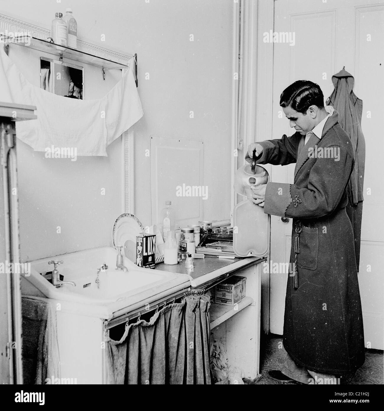 1950s, un immigrato d'oltremare appena arrivato in Gran Bretagna in piedi al lavabo nel suo letto, versando acqua da un bollitore in una bottiglia di acqua calda. Foto Stock