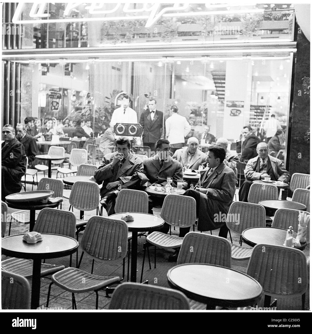 1950s. Presto la sera e giovani uomini parigino seduti in sedie di vimini gustando un drink e discussione insieme al di fuori di un cafe bar, Parigi, Francia. Foto Stock 1950s. Presto la sera e giovani uomini parigino seduti in sedie di vimini gustando un drink e discussione insieme al di fuori di un cafe bar, Parigi, Francia. Foto Stock