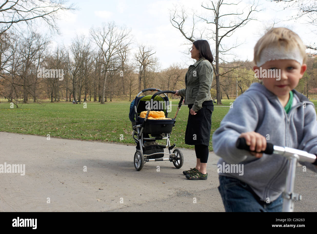 Un giovane bambino biondo ragazzo su uno scooter nel parco con testa di feriti (capeline bendaggio) senza casco di sicurezza. Madre con la PRAM indietro Foto Stock