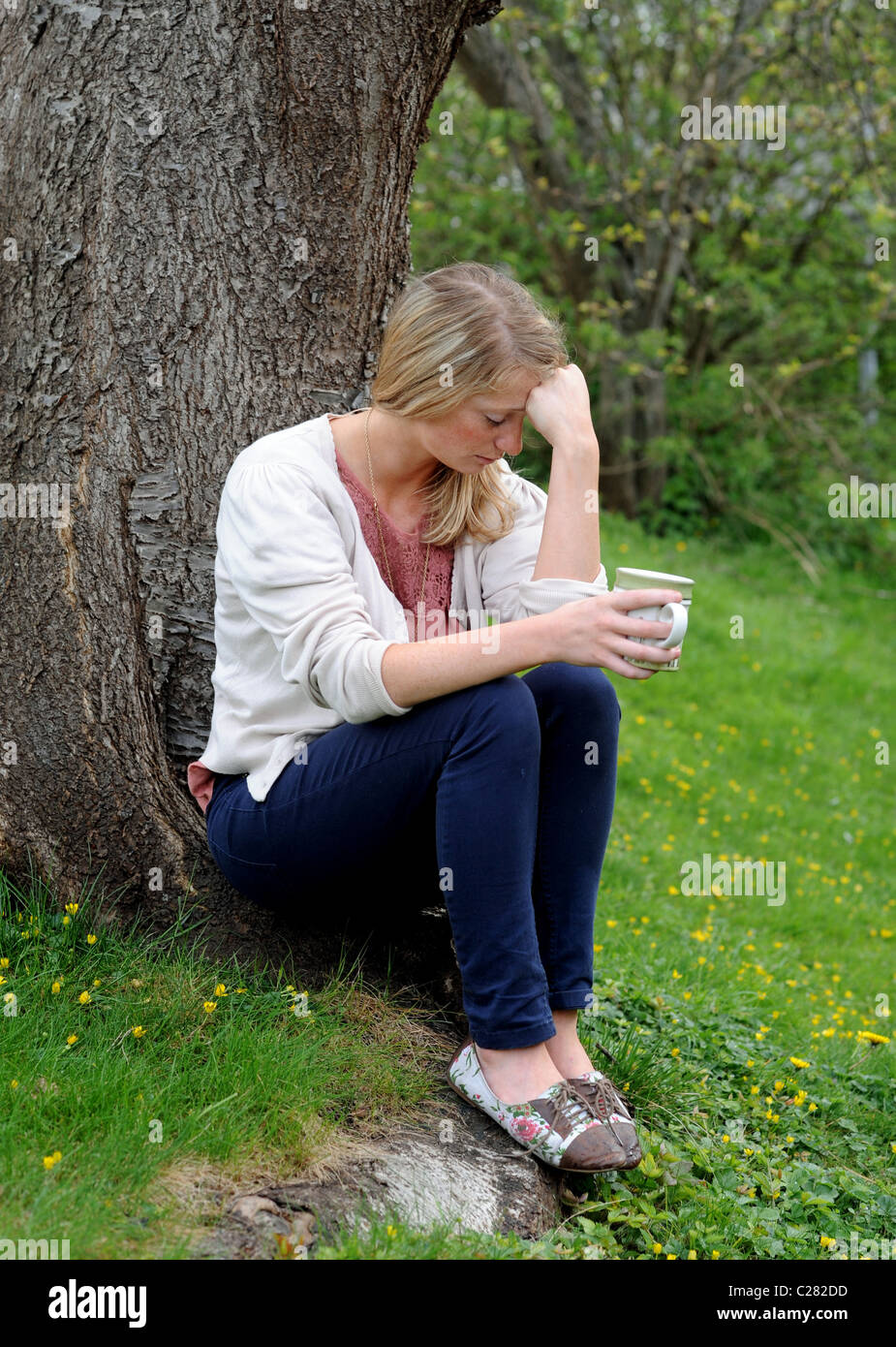 Giovane donna nella disperazione si siede da un albero con una tazza di tè in testa le mani molto turbato - poste da modello a modello Foto Stock