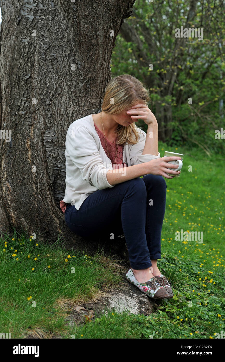 Giovane donna nella disperazione si siede da un albero con una tazza di tè in testa le mani molto turbato - poste da modello a modello Foto Stock