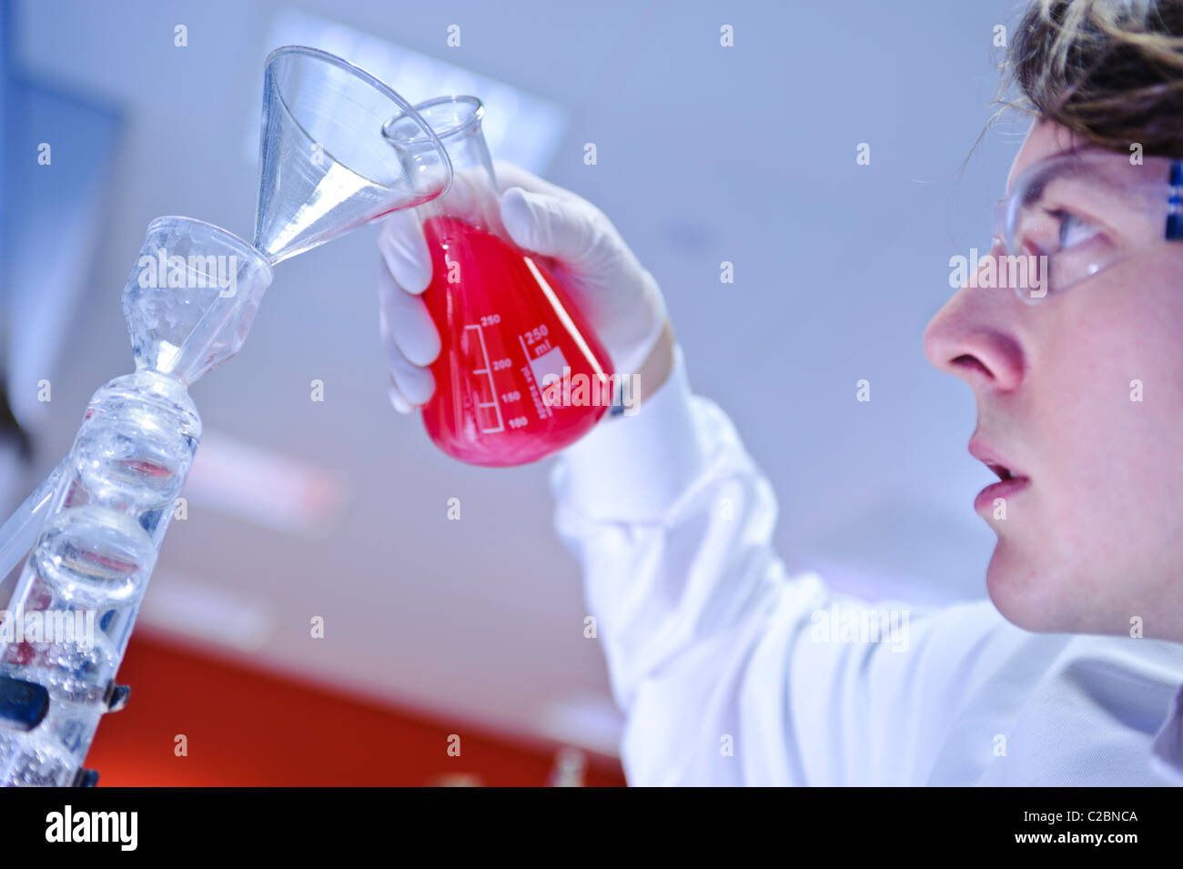 I giovani fanno scienziato degli occhiali guanti e camice versando rosso brillante liquido dal bicchiere di vetro nel laboratorio di scienze Foto Stock