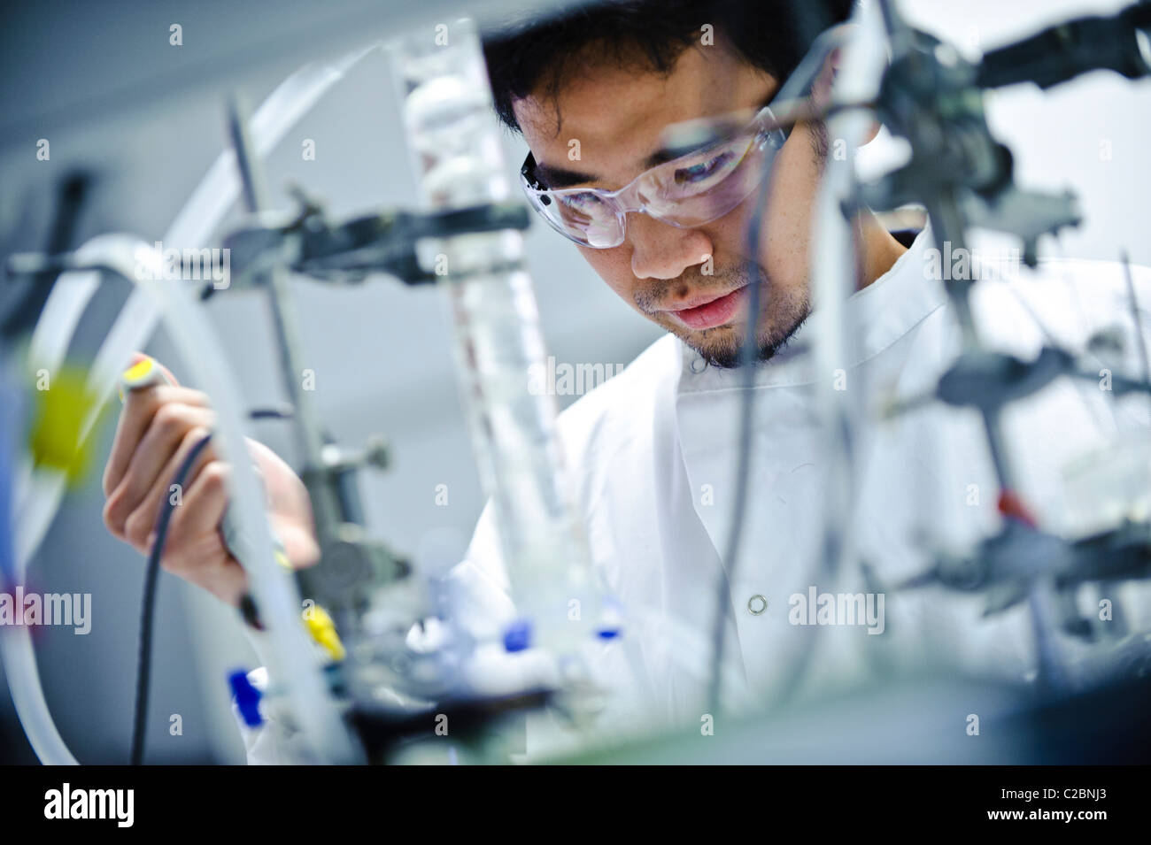 Giovani asiatici scienziato maschio indossando occhiali protettivi trasparenti di colore bianco e la scienza ricoprire guardando il tubo di vetro della apparecchiatura in laboratorio di scienze Foto Stock