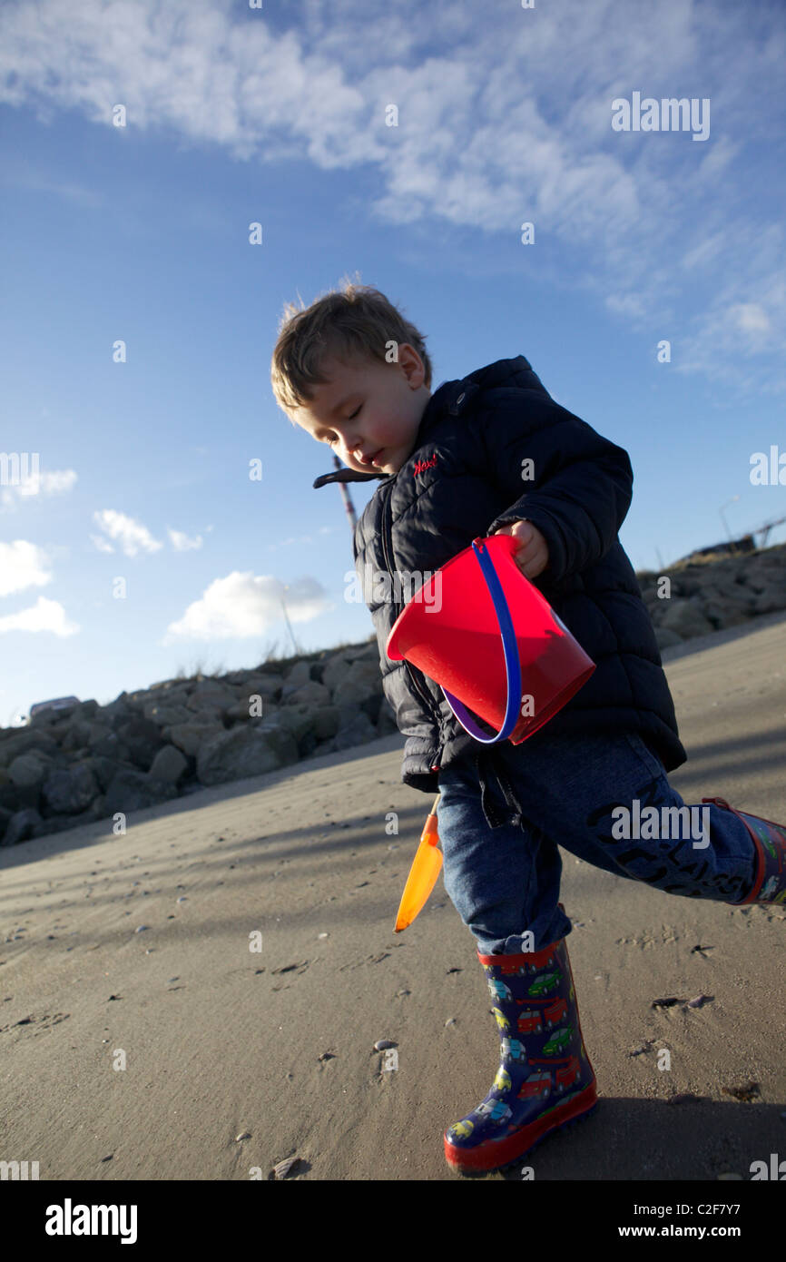 Piccolo Ragazzo con una benna di rosso e giallo beachcombing a forcella e alla ricerca di conchiglie Foto Stock