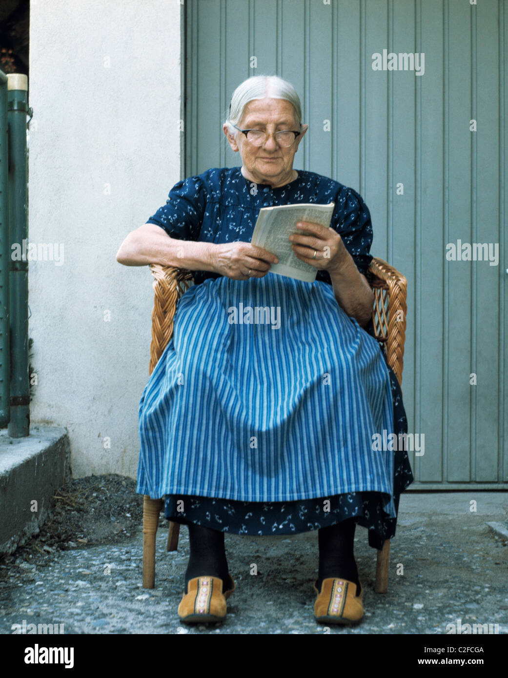 Negli anni settanta, la gente, la vecchiaia, la donna più anziana seduti in  poltrona a leggere un libro a piena lunghezza ritratto, grembiule,  abbigliamento, calzature da casa, zoccoli, pantofole, di età compresa