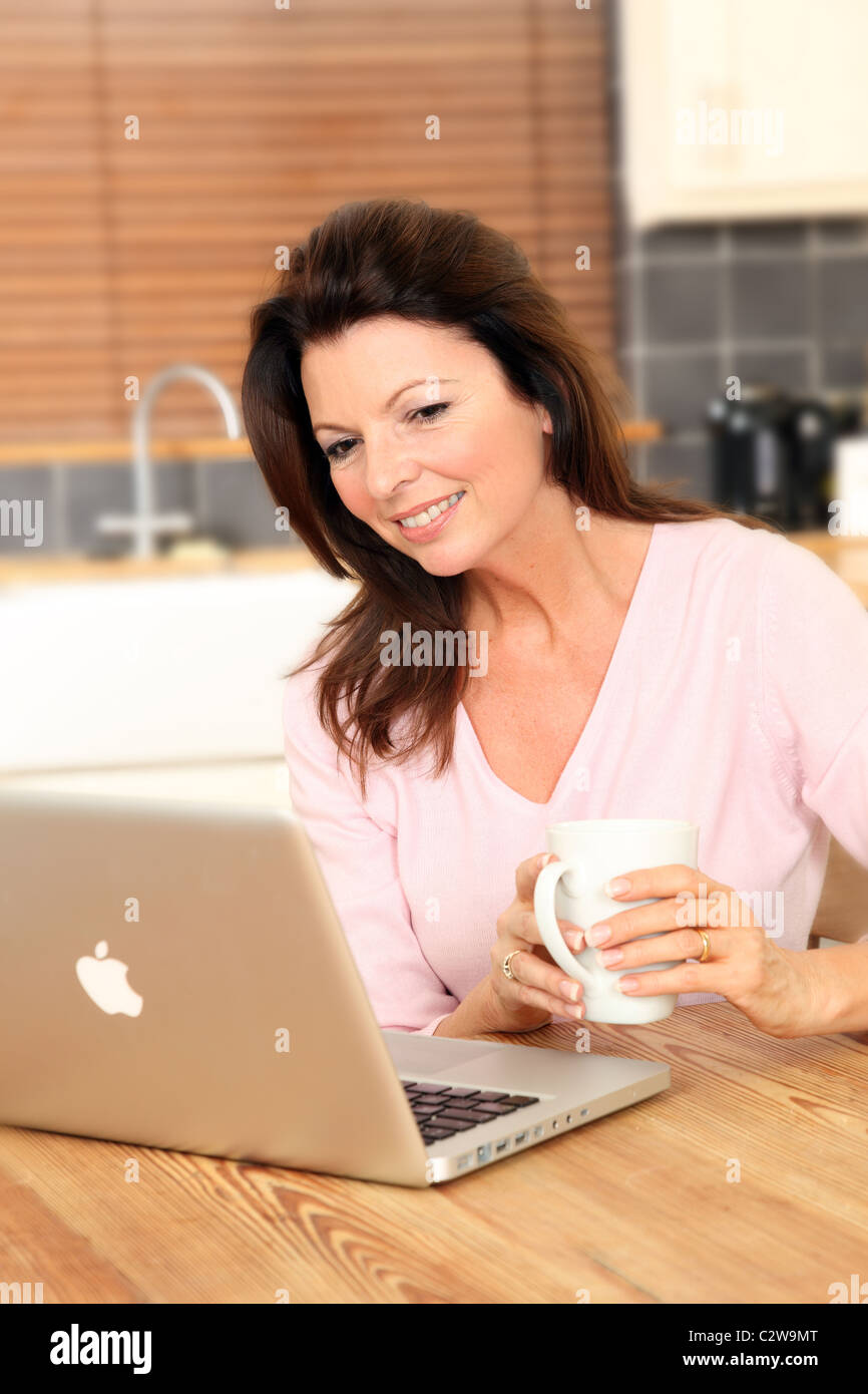 Donna che utilizza il suo computer portatile in cucina tenendo una tazza di tè o caffè Foto Stock