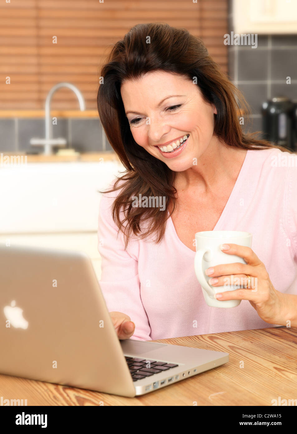 Donna felice con il suo computer portatile in cucina tenendo una tazza di tè o caffè Foto Stock