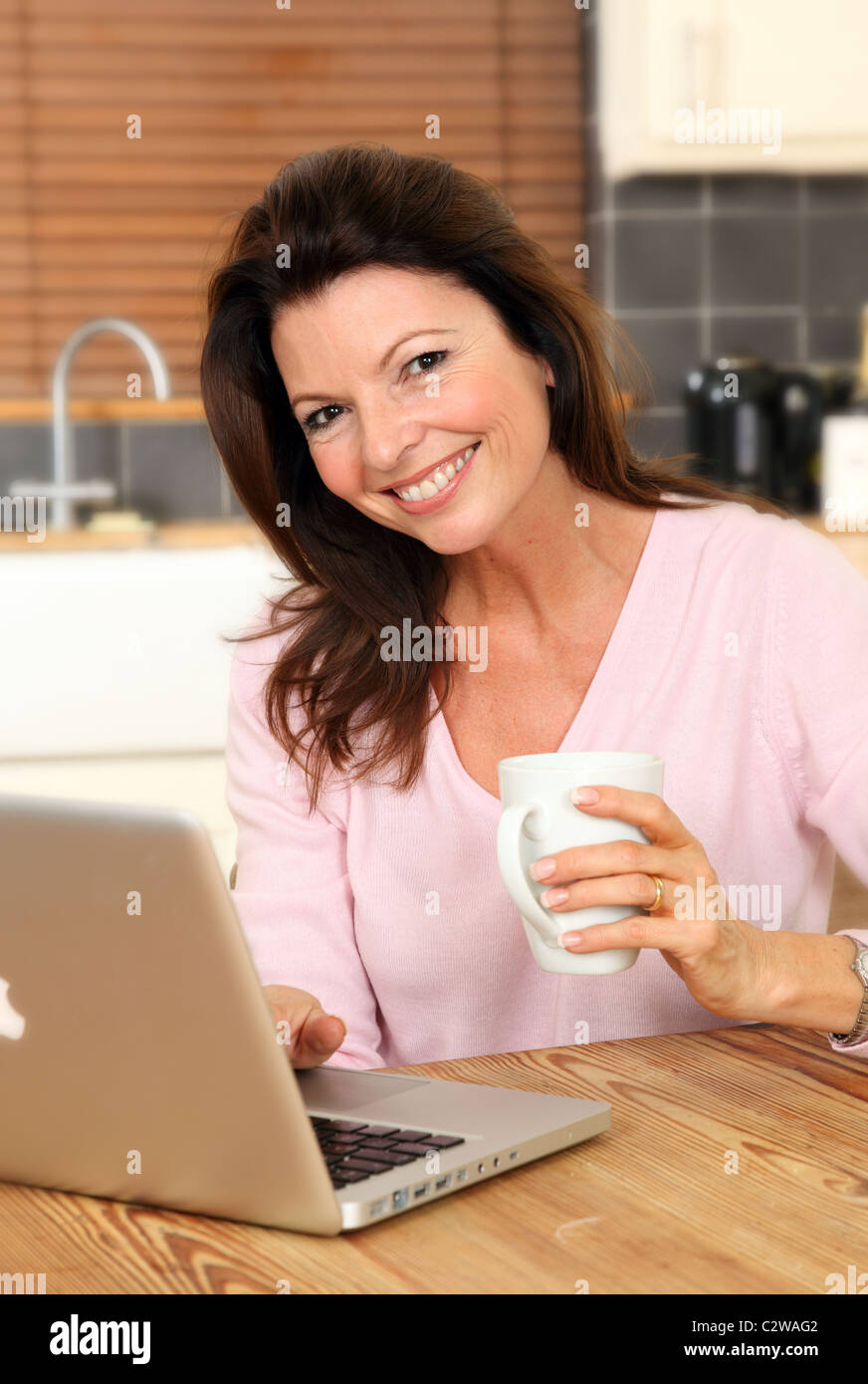 Donna felice con il suo computer portatile in cucina tenendo una tazza di tè o caffè, guardando la fotocamera Foto Stock
