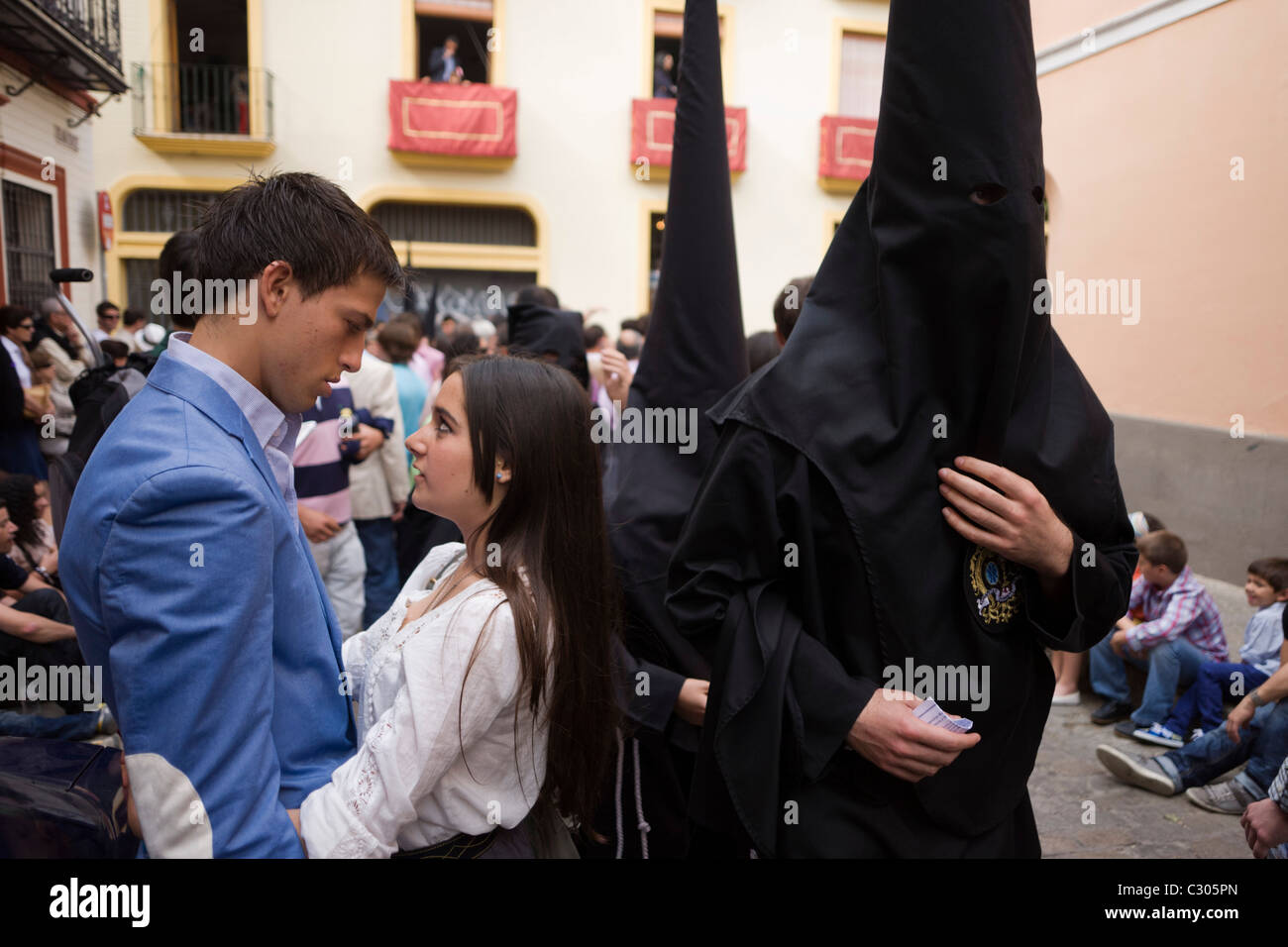 I penitenti incappucciati (Nazarenos) a piedi passato giovani innamorati durante il Siviglia annuale della pasqua settimana santa (Semana Santa de Sevilla) Foto Stock