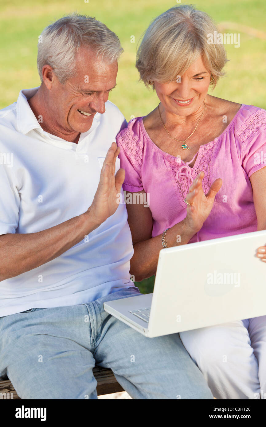 Felice senior uomo & donna giovane seduto insieme al di fuori nel sole sventolare mentre si utilizza un computer per internet VOIP chiamata telefonica Foto Stock