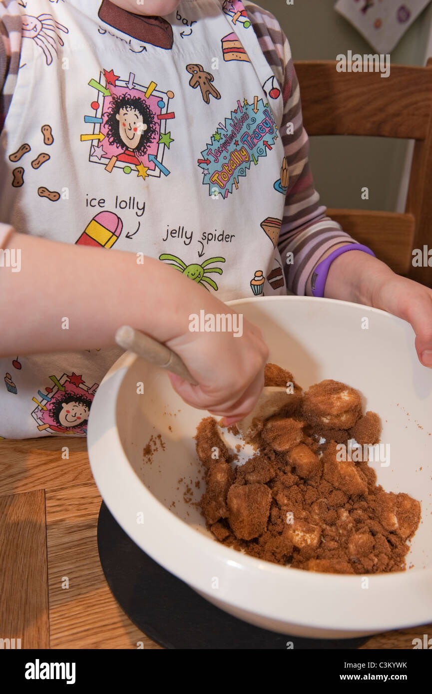Torta di cottura della ragazza giovane, mescolando gli ingredienti (burro & zucchero di canna) in ciotola con il cucchiaio di legno & che indossa il grembiule del Beaker di Tracy - Yorkshire, Inghilterra UK Foto Stock