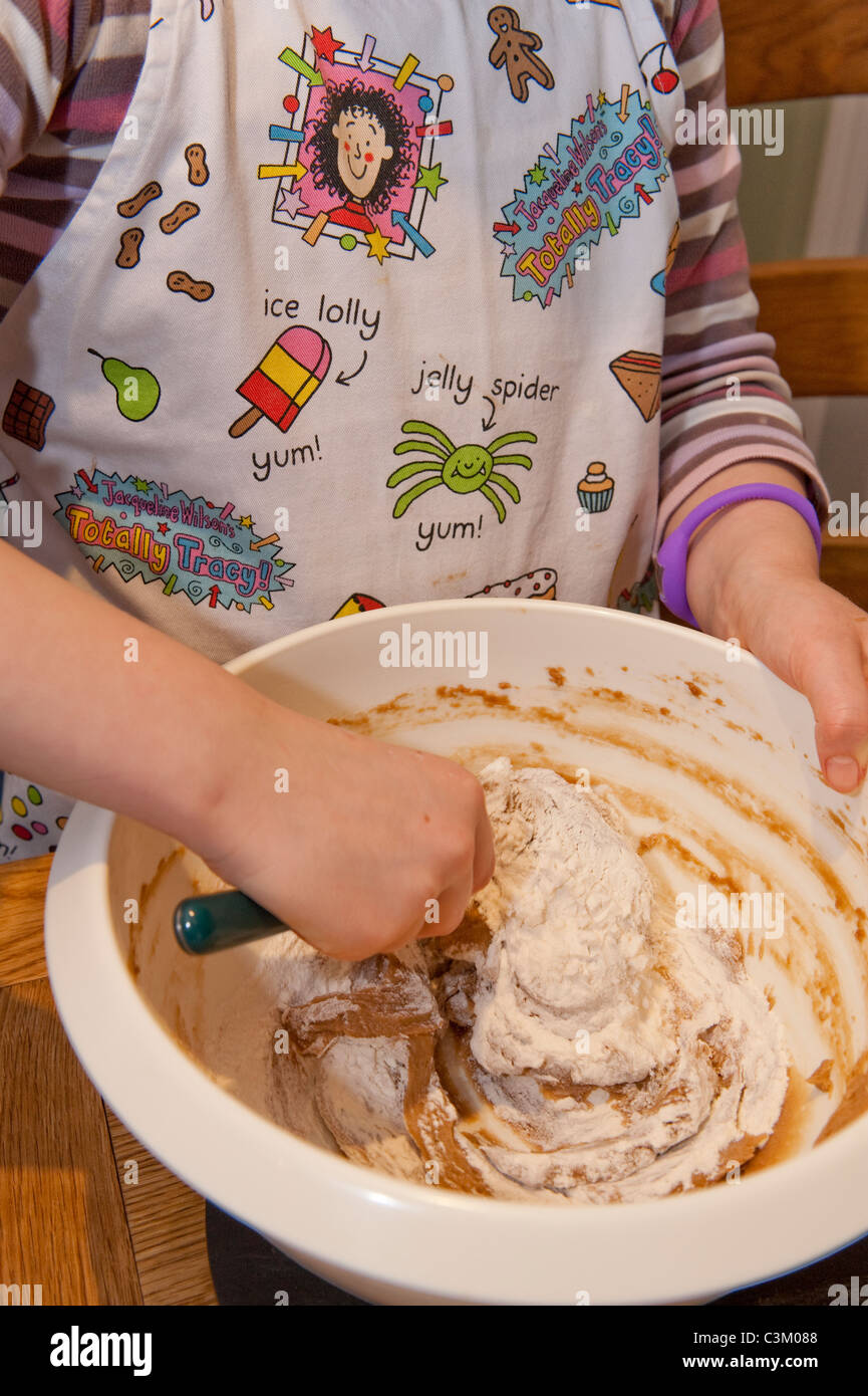 Giovane ragazza che cuoce torta al cioccolato, piegando la farina in miscela (primo piano di mani, miscela, manico di cucchiaio e grembiule di Tracy Beaker) - Yorkshire, Inghilterra, UK. Foto Stock