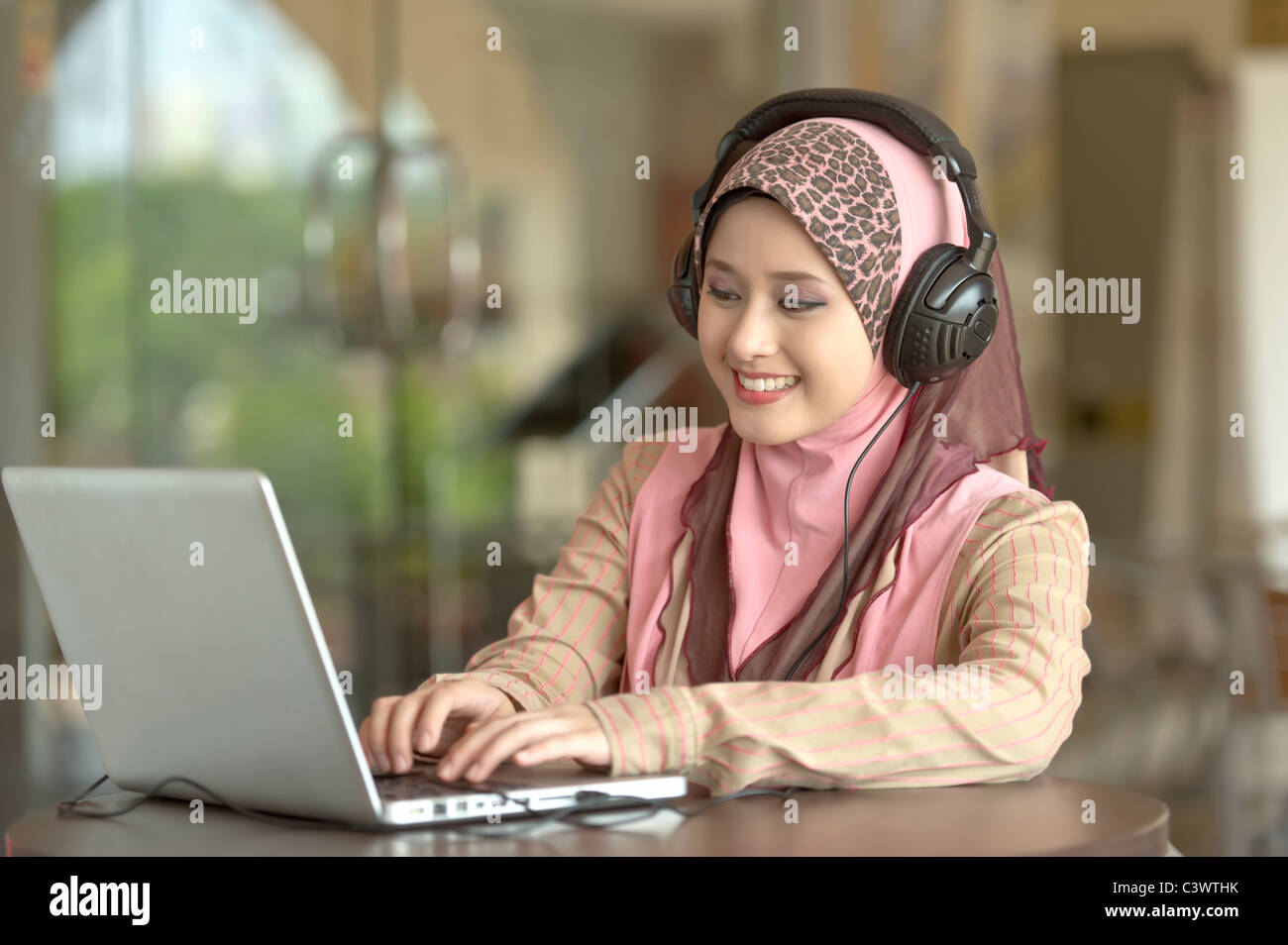 Giovani asiatici bella donna musulmana in testa sciarpa ascolta audio con testa il telefono mentre si lavora sul computer portatile in cafe Foto Stock
