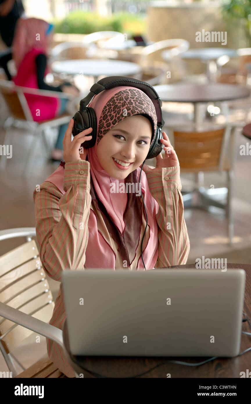 Giovani asiatici bella donna musulmana in testa sciarpa ascolta audio con le cuffie mentre si lavora sul computer portatile in cafe Foto Stock