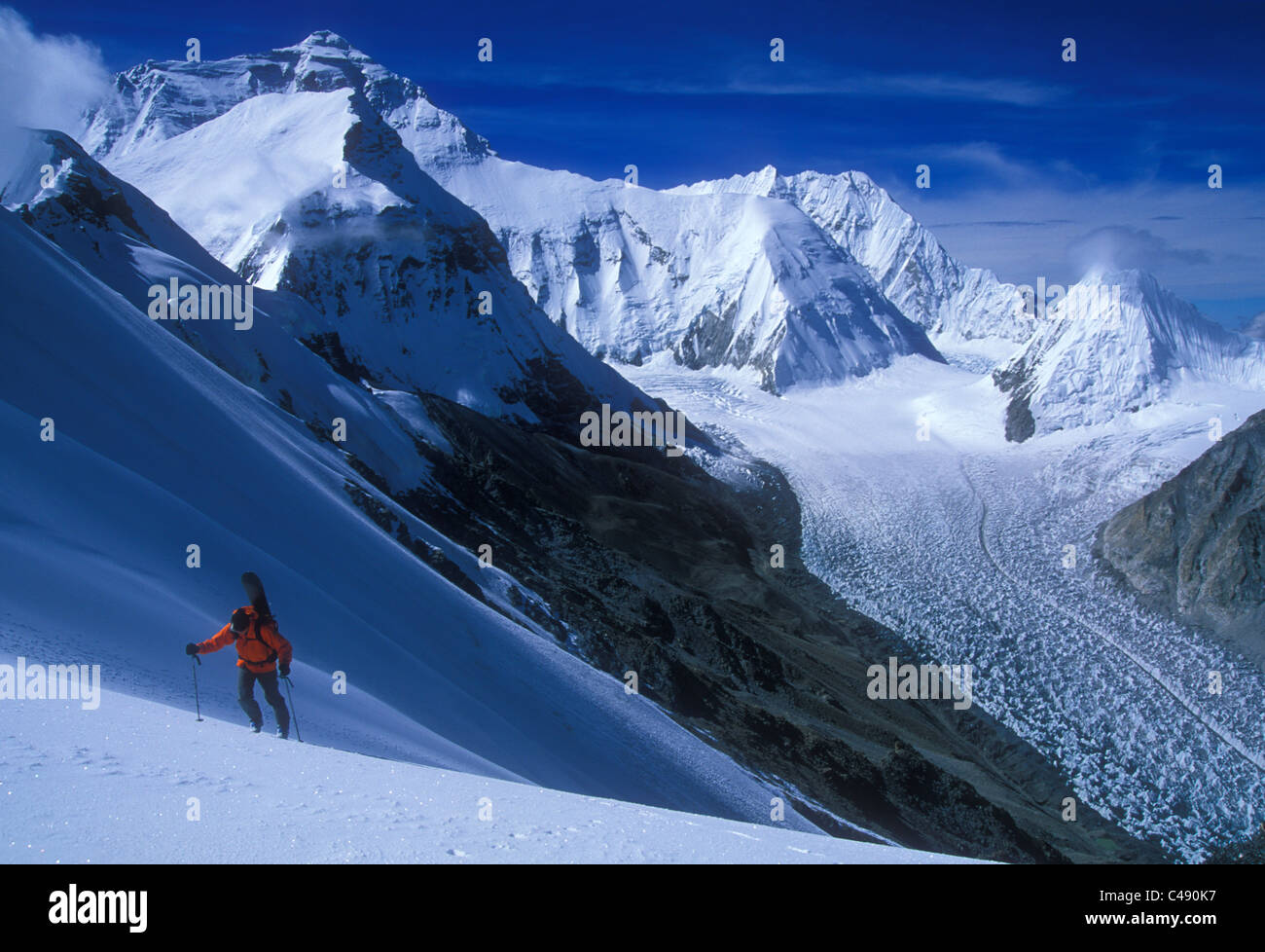 Un uomo che porta il suo snowboard si arrampica su di una collina con un ghiacciaio in background. Foto Stock