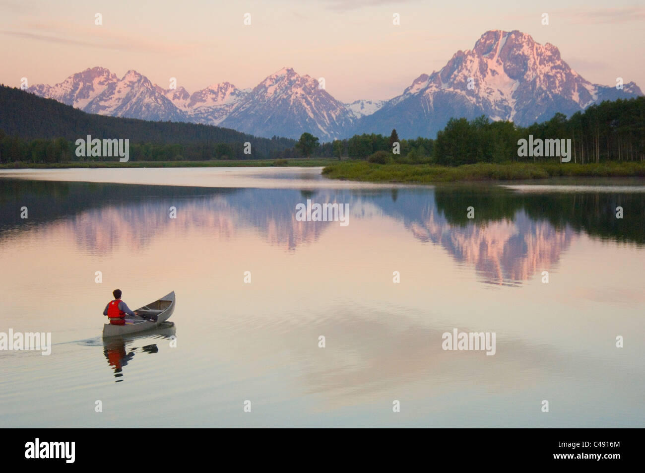 Un uomo in canoa sul fiume calmo presso sunrise con enormi montagne coperte di neve in background. Foto Stock