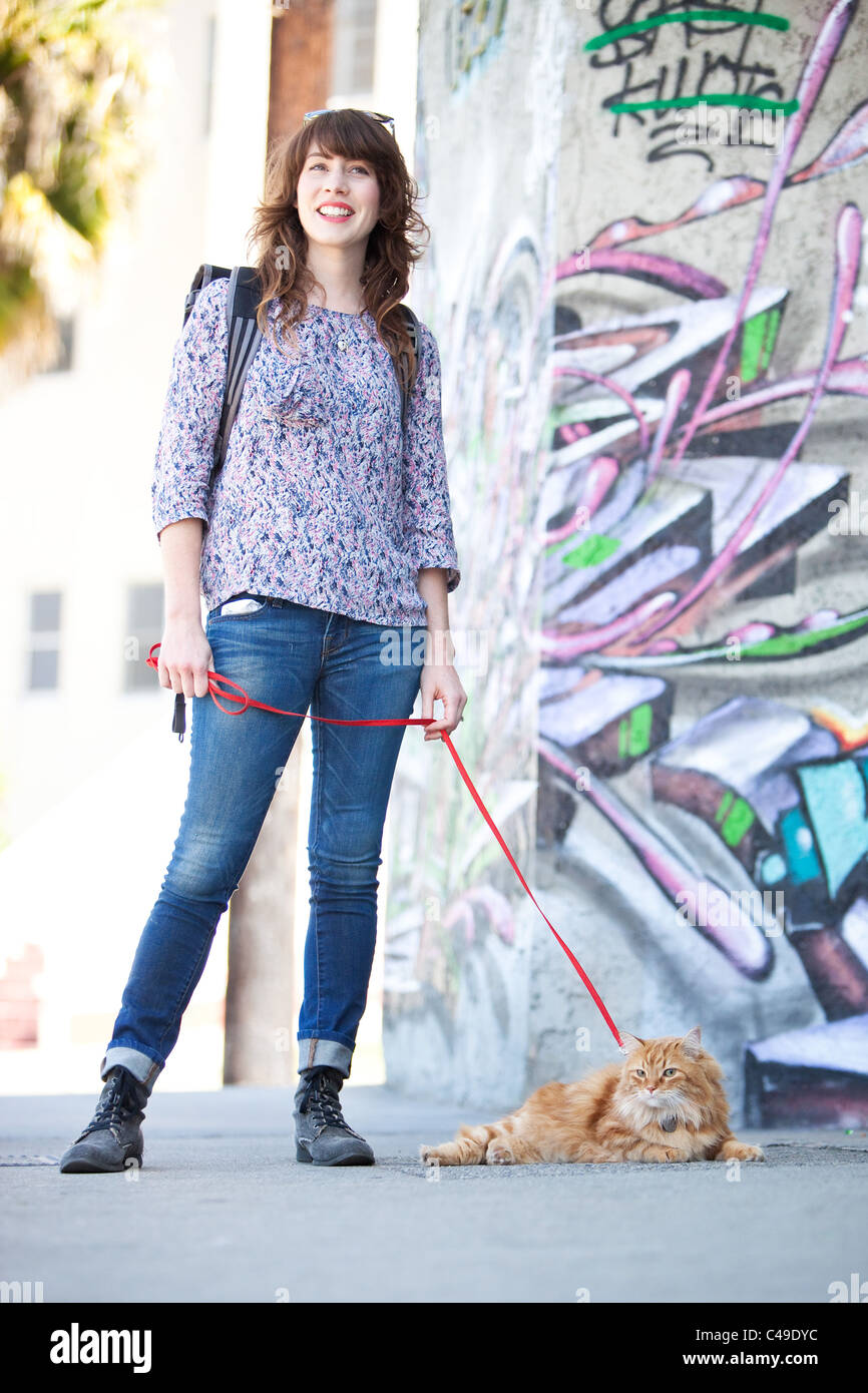 Un sorridente giovane donna con un arancione longhaired Manx cat al guinzaglio, stando in piedi in un'area urbana con graffiti. Foto Stock