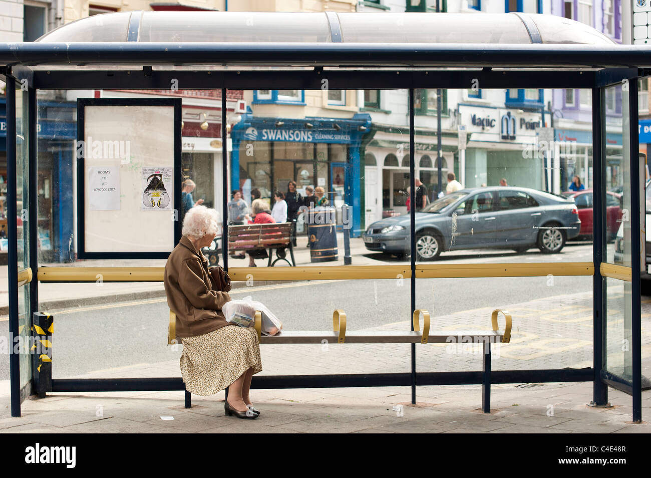 Un anziano senior donna seduta da sola in un bus shelter in attesa per i mezzi di trasporto pubblici, REGNO UNITO Foto Stock
