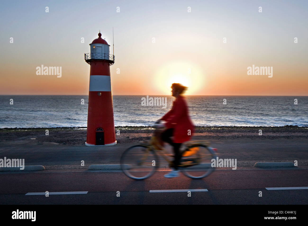 Donna Bicicletta Equitazione lungo faro rosso al tramonto, Westkapelle, Zeeland, Paesi Bassi Foto Stock