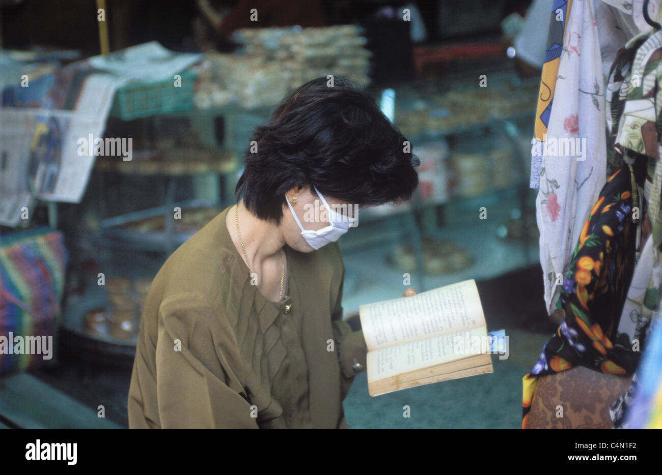 Donna thai trader, indossando un anti-inquinamento maschera, la lettura di un libro in un negozio di Bangkok Foto Stock