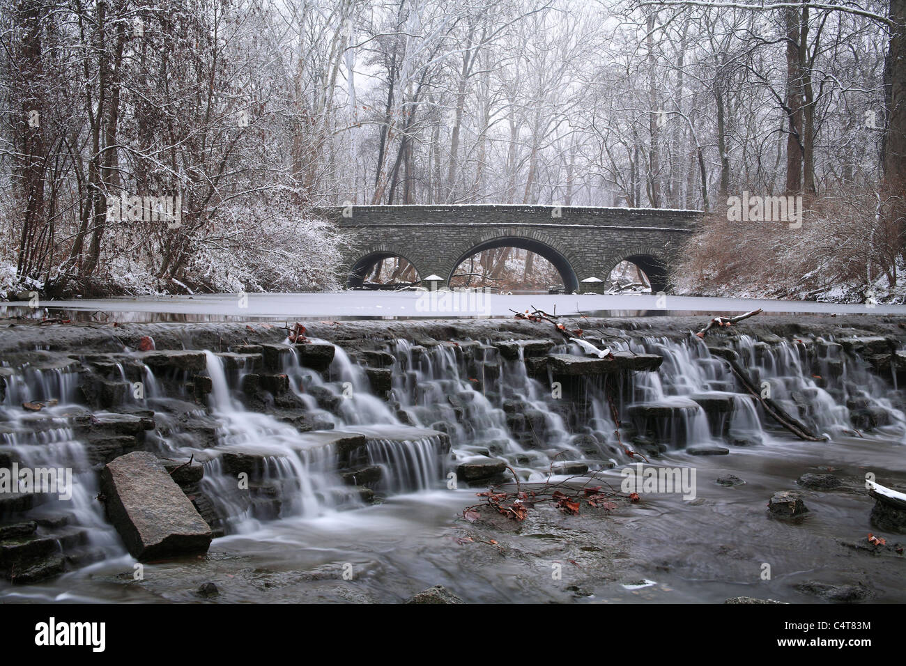 Coperta di neve alberi che incornicia un ponte di pietra e cascata durante l'Inverno nel parco, Sharon boschi, Southwestern Ohio, Stati Uniti d'America Foto Stock