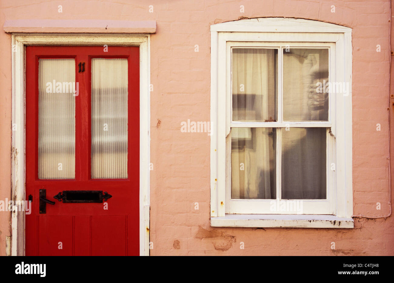 Dettaglio della casa rosa con pareti dipinte di rosso porta anteriore e sash bianco finestra con tendina inondata dalla calda luce del sole riflessa Foto Stock