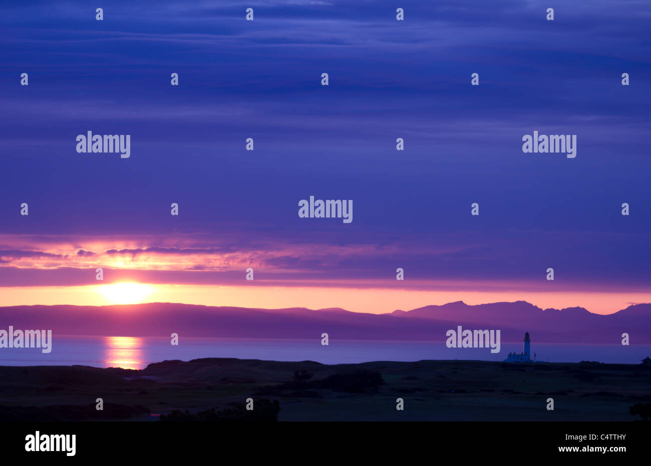 Il sole che tramonta su un faro con le montagne sullo sfondo Foto Stock