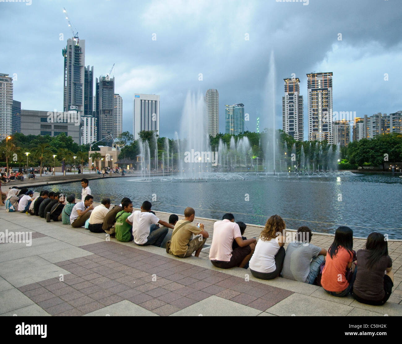 La gente seduta su una scalinata di KLCC Park presso le Petronas Twin Towers di fronte alla skyline con edifici per uffici e alberghi Foto Stock