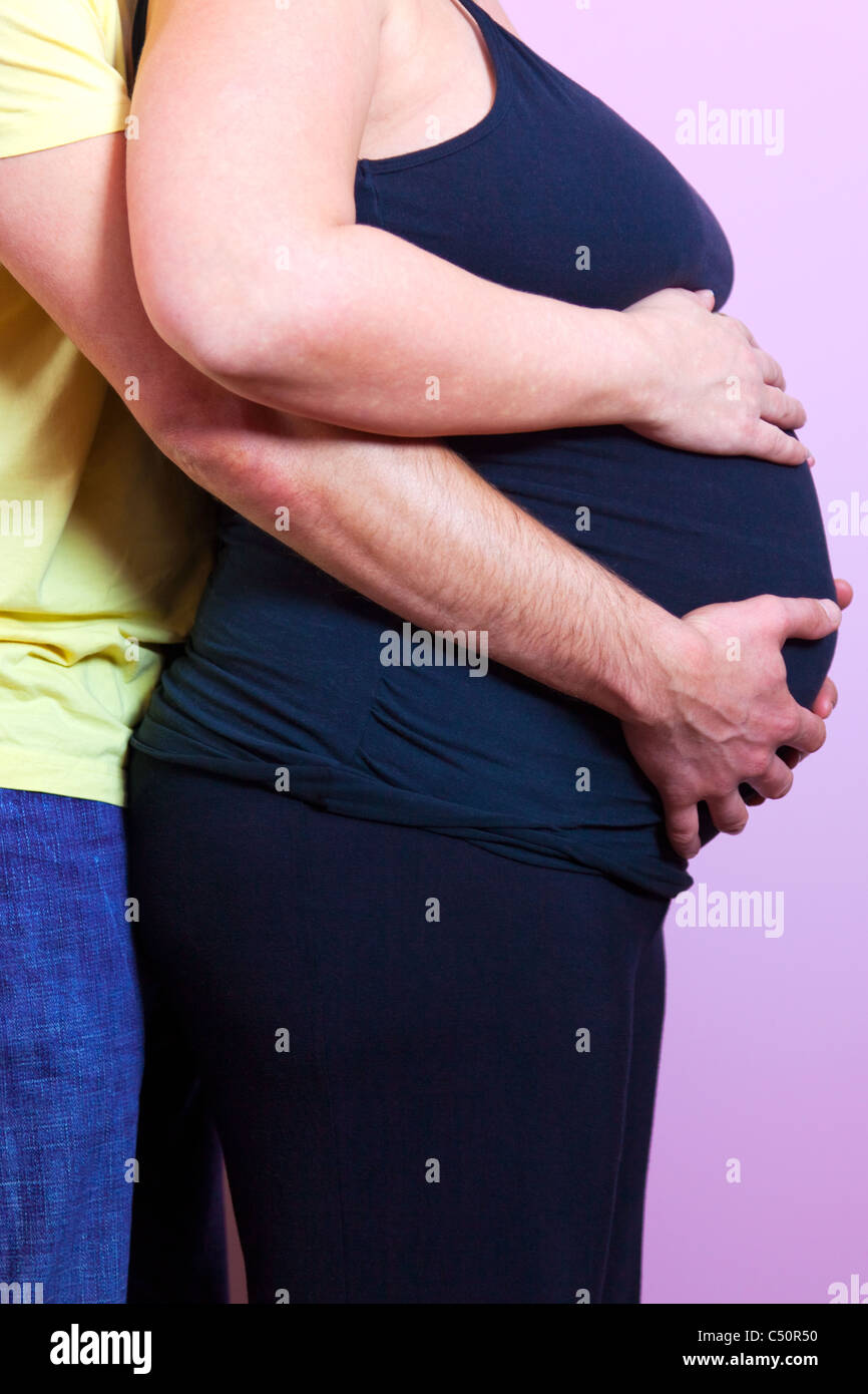 Foto di una donna incinta con il marito dietro entrambi con le loro mani sul suo stomaco Foto Stock