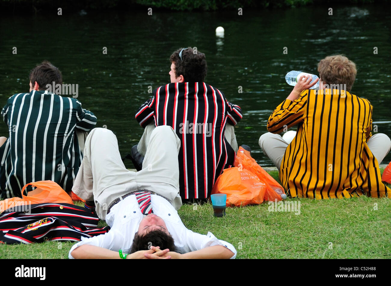 I ragazzi in blazer a strisce seduti lungo il bordo del fiume Tamigi durante Henley Royal Regatta, Henley on Thames, Oxfordshire, Regno Unito Foto Stock