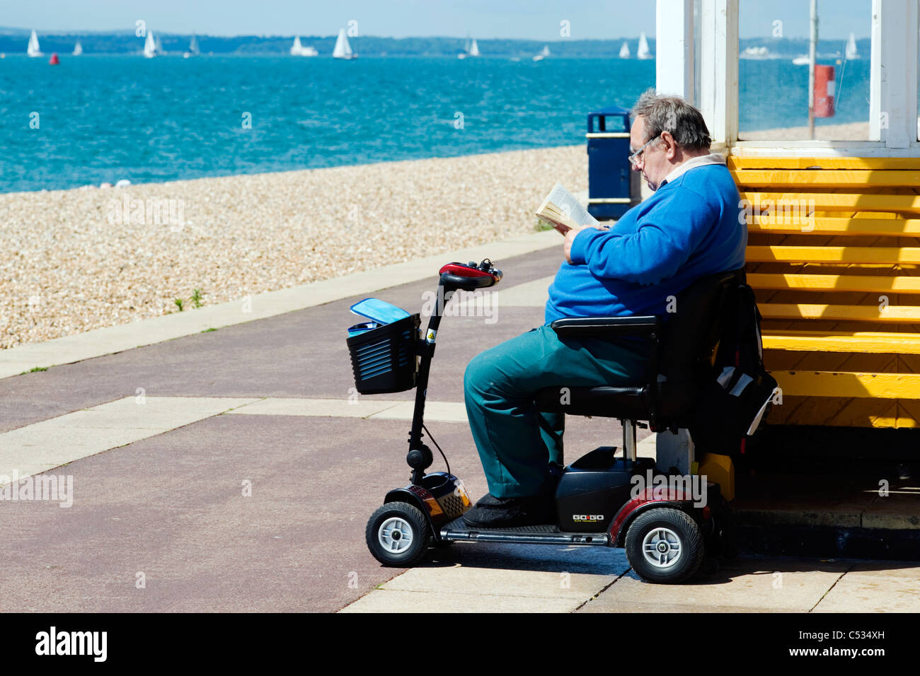 L uomo sulla mobilità scooter la lettura di un libro su Southsea seafront Foto Stock