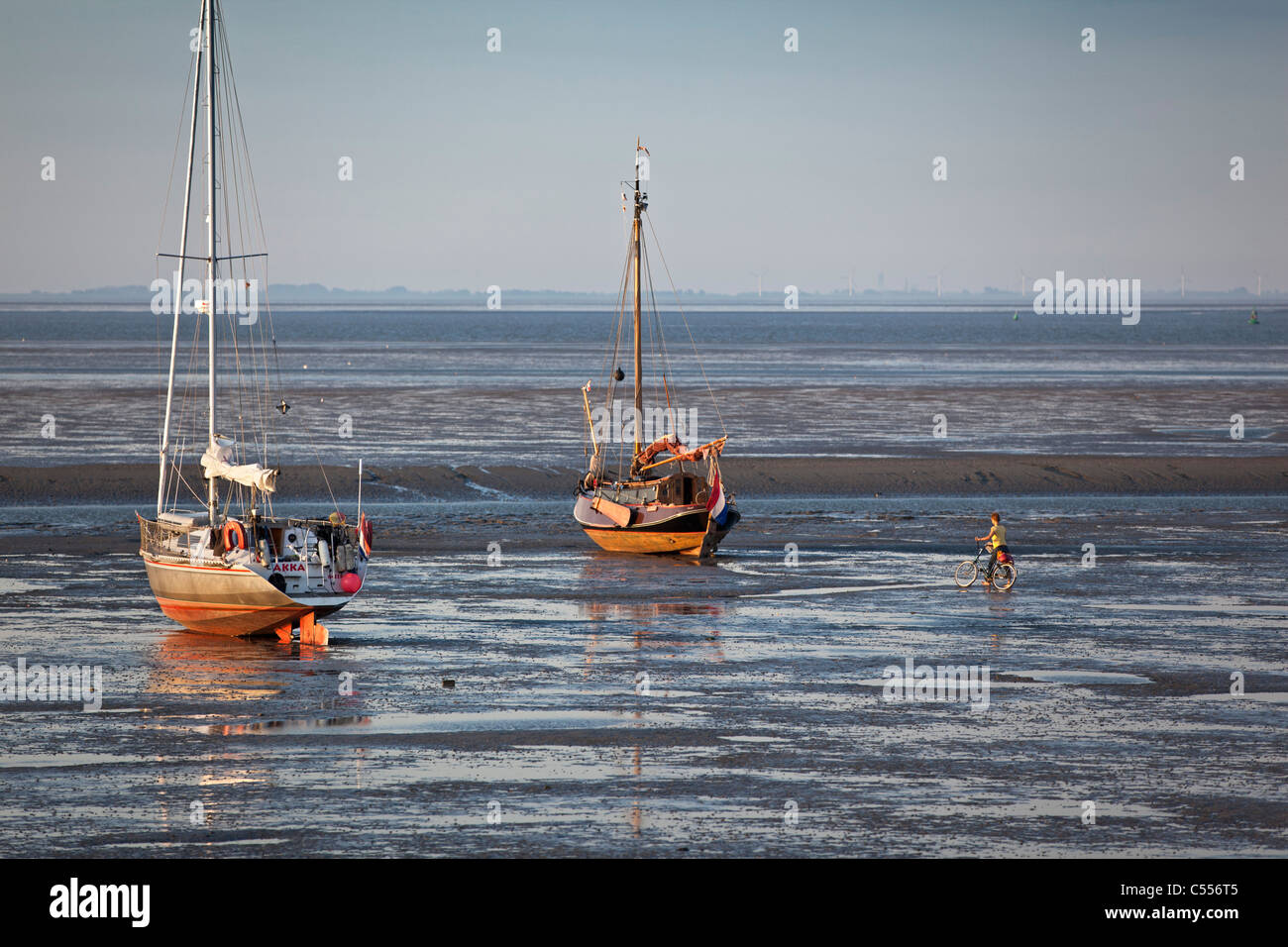I Paesi Bassi, il Nes Ameland, Isola, appartenente al mare di Wadden Islands. Barche a vela sulla piana di fango nel porto. Donna e bicicletta. Foto Stock