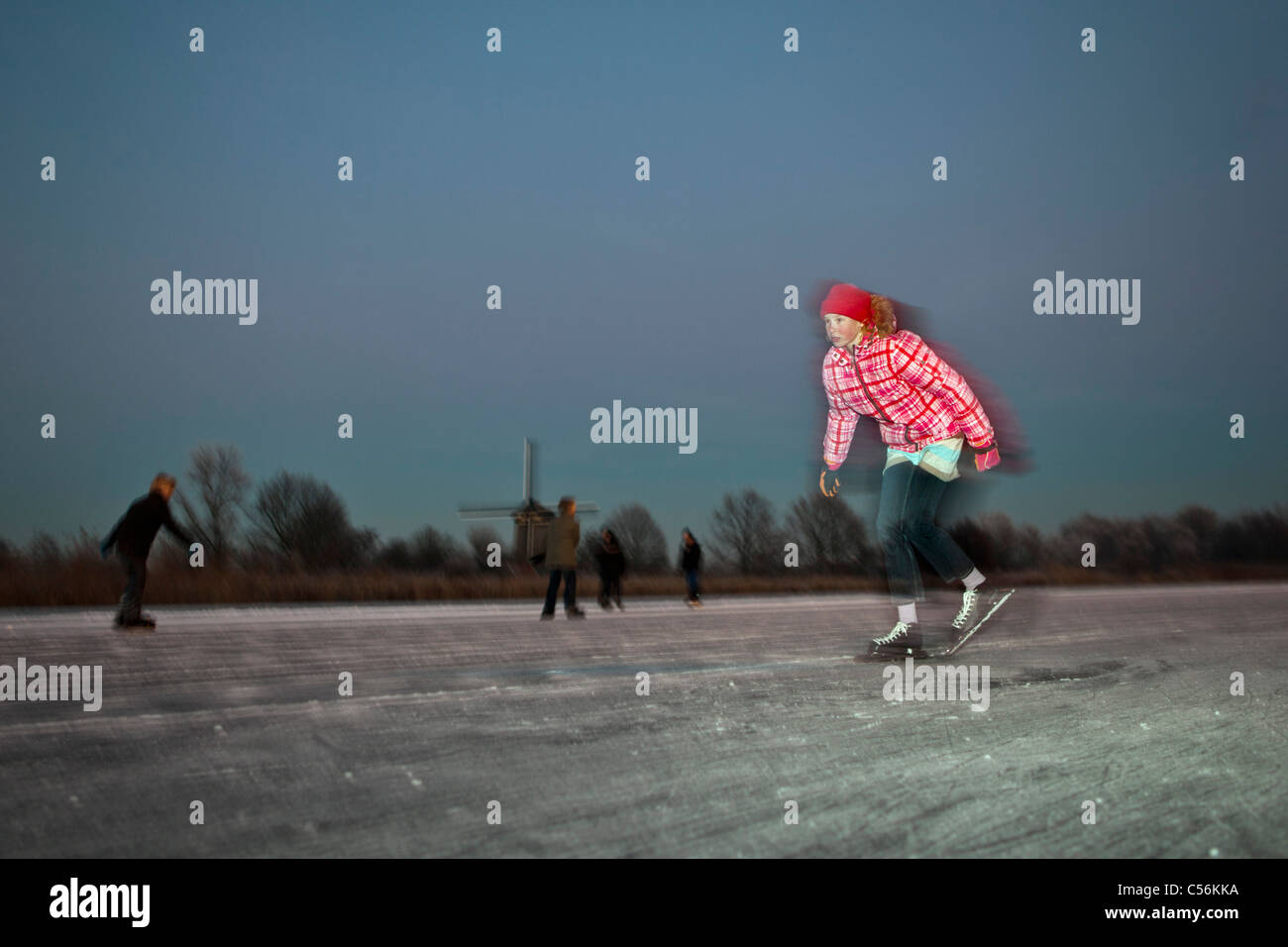 I Paesi Bassi, Ankeveen. Ragazza il pattinaggio su ghiaccio. Il mulino a vento di sfondo. Crepuscolo. Motion sfocata. Foto Stock