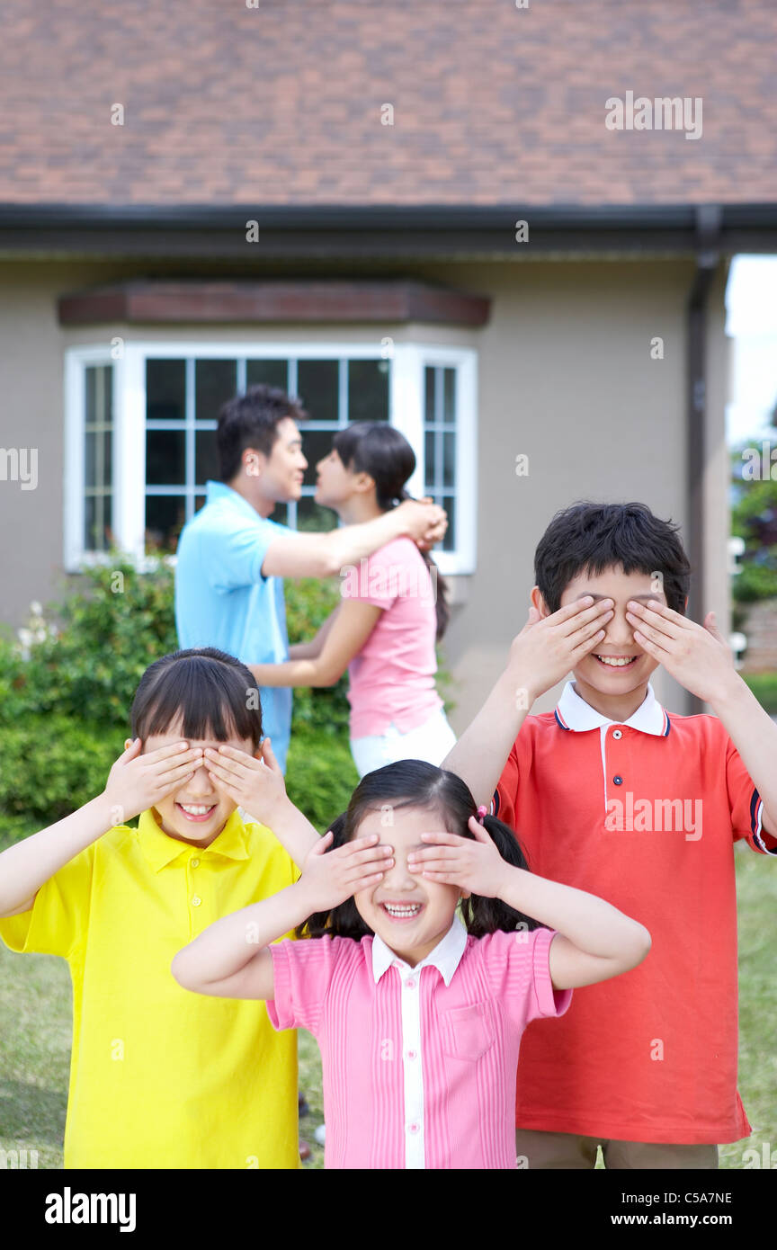 I bambini si tengono per mano sugli occhi, mentre i genitori baciare in background Foto Stock
