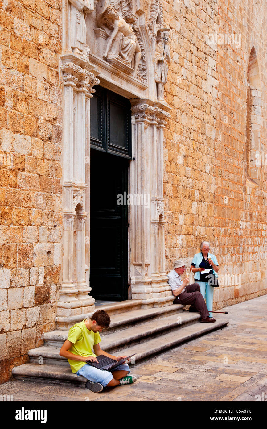 Giovane uomo su un computer portatile e la coppia di anziani seduti sui gradini della vecchia chiesa edificio a Dubrovnik, Dalmazia Croazia Foto Stock