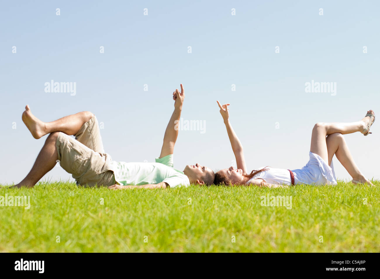 Coppia giovane sdraiarsi sull'erba e punto verso il cielo, per esterno in posizione di parcheggio Foto Stock