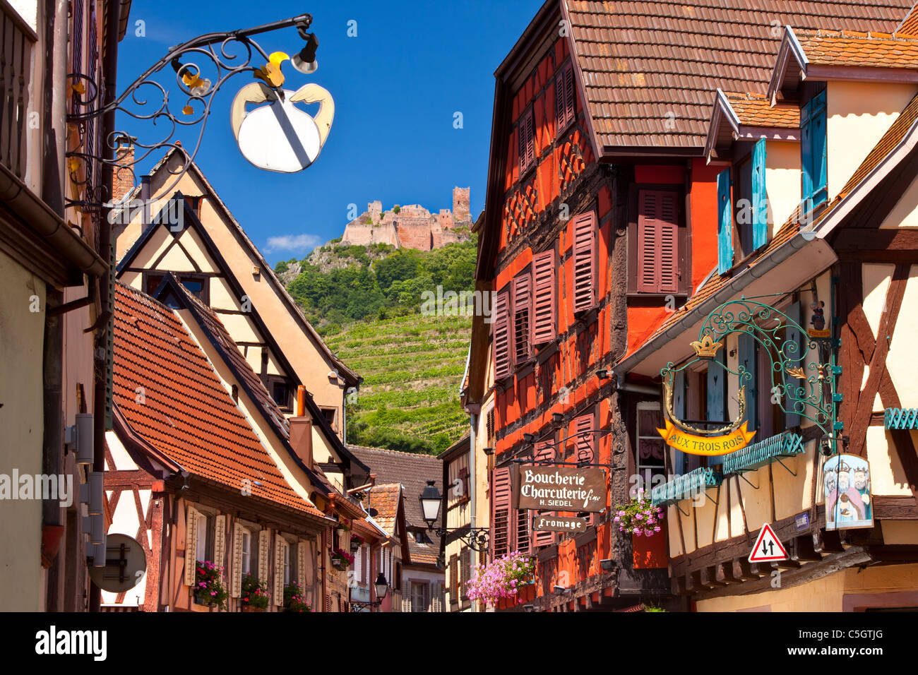 Negozi e abitazioni lungo la Grand Rue con le rovine del Castello di San Ulrico oltre, Ribeauville, Alsazia Haut-Rhin Francia Foto Stock