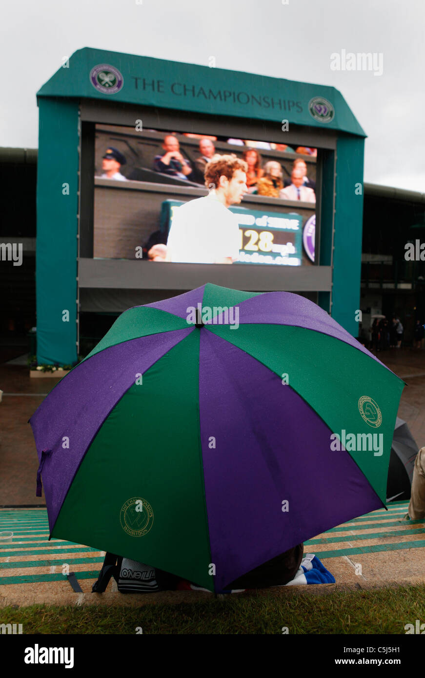 Spettatore sotto un ombrello guardando giocare sul grande schermo a Wimbledon Foto Stock