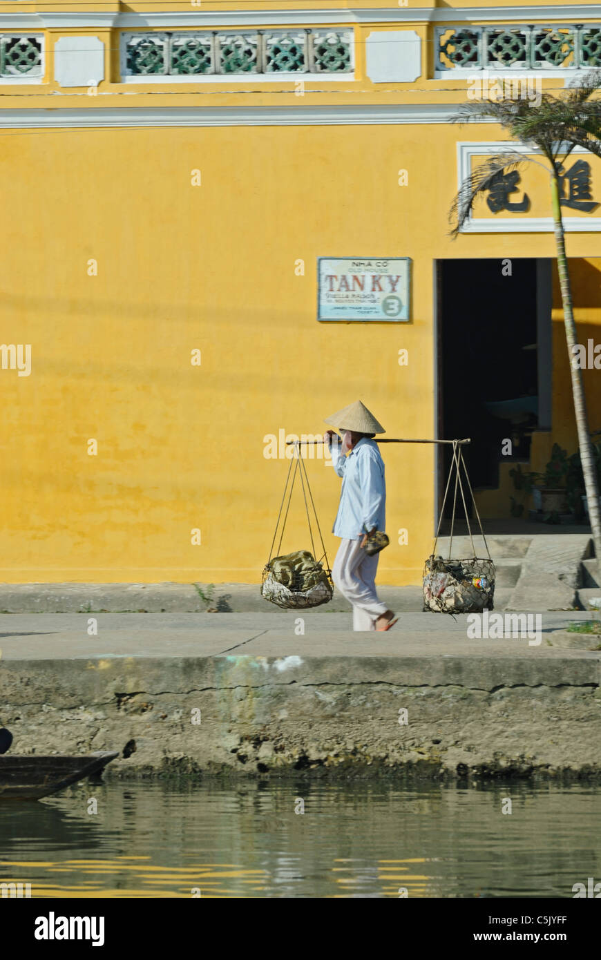 Asia, Vietnam, Hoi An. Hoi An old quarter. Donna vietnamita che trasportano merci lungo la Bach Dang passeggiata sul fiume. Il centro storico Foto Stock