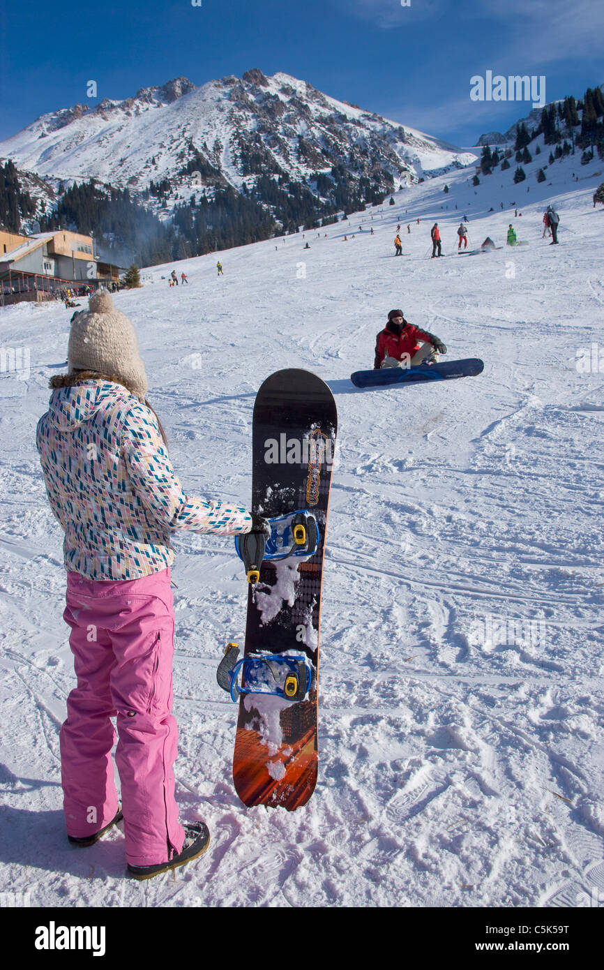 Ragazza con lo snowboard guardando un altro caduto snowboarder, snowy ski resort di Chimbulak, Kazakistan Foto Stock